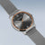 Herrenarmbanduhr schräg: graues Textilband, dunkles Zifferblatt, orangefarbene Lünette