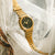Goldfarbene Armbanduhr mit schwarzem Zifferblatt und perforiertem Metallarmband.